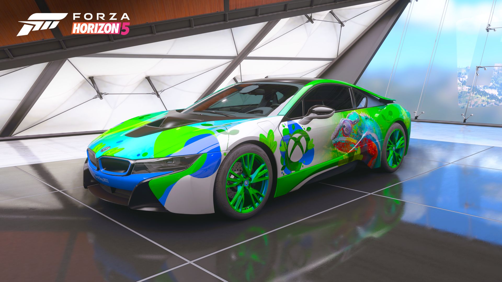 Ein 2015er BMW i8 steht in einer professionell beleuchteten Garage. Das Auto ist in Blau-, Grün- und Weißtönen lackiert und weist Formen auf, die an Pflanzenlaub erinnern. Das Xbox-Logo ist an der Seite des Fahrzeugs zu sehen, neben einem detaillierten Bild eines Chamäleons in ähnlichen Farben.