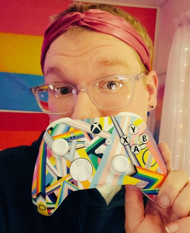 ALT TEXT: ItsMileyGirl hält sich einen Xbox Pride-Controller vor den Mund, mit einer Pride-Flagge im Hintergrund. Sie trägt eine durchsichtige Brille und ein rosa Stirnband.