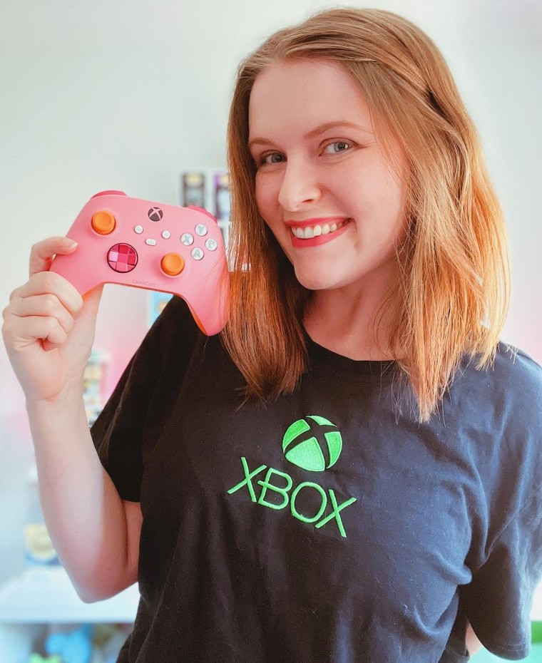 ALT TEXT: ctrlaltcraft lächelt in einem Xbox T-Shirt und hält einen rosa-orangen Xbox-Controller neben ihr Gesicht.