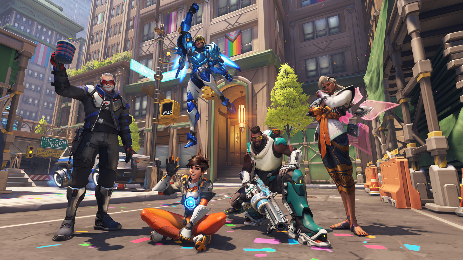 Wide shot of the Overwatch 2's Midtown map, customized with rainbow-colored theme and Pride flags and from left to right, Soldier 76 raising a cup, Tracer waving while sitting in criss-cross, Pharah celebrating mid-air, Baptiste crouching and Lifeweaver posing for the picture.