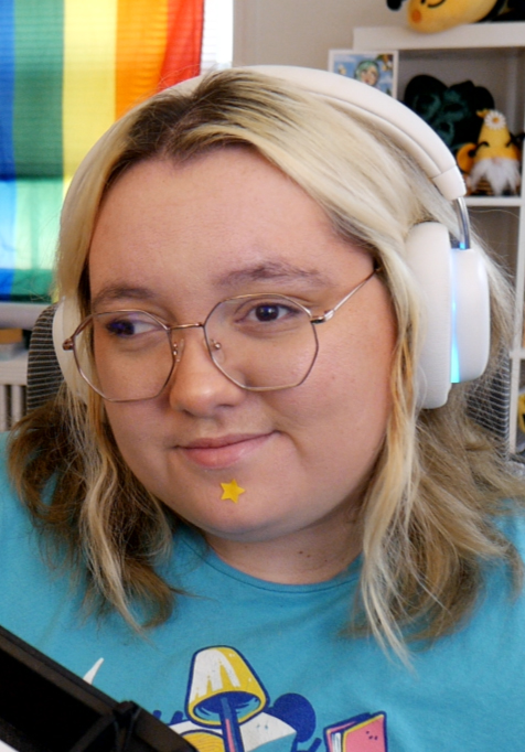 Queerlybee smiles at the camera wearing a blue tshirt and white headphones. A rainbow Pride flag is displayed behind them.