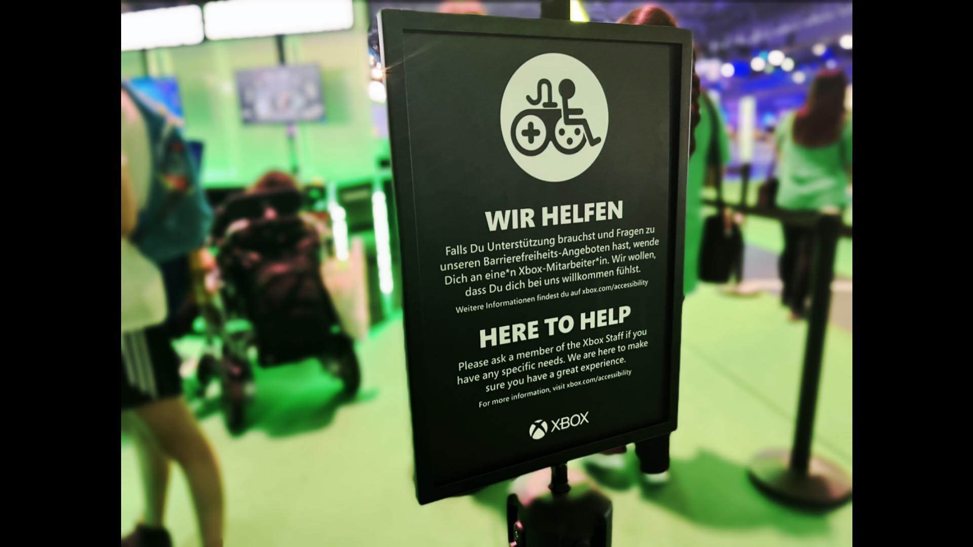 A black sign with a white gaming accessibility logo stands in front of demo stations at a game convention. Text reads in English and German, "Here to help. Please ask a member of the Xbox Staff if you have any specific needs. We are here to make sure you have a great experience. For more information, visit xbox.com/accessibility. Xbox."